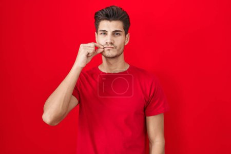 Foto de Joven hombre hispano de pie sobre fondo rojo boca y labios cerrados como cremallera con los dedos. secreto y silencioso, tabú hablando - Imagen libre de derechos