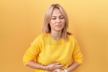 Téléchargez les photos : Jeune femme caucasienne portant un pull jaune avec la main sur le ventre parce que l'indigestion, la maladie douloureuse se sent mal. concept de douleur. - en image libre de droit