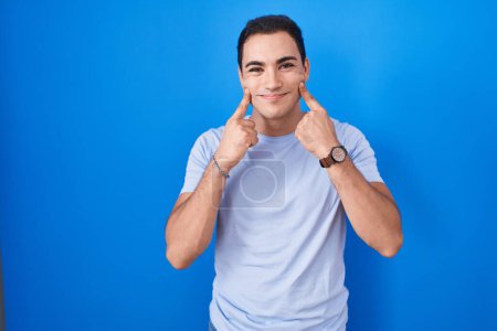 Téléchargez les photos : Young hispanic man standing over blue background smiling with open mouth, fingers pointing and forcing cheerful smile - en image libre de droit