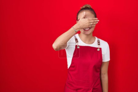 Foto de Mujer hispana joven vistiendo delantal de camarera sobre fondo rojo sonriendo y riendo con la mano en la cara cubriendo los ojos para sorpresa. concepto ciego. - Imagen libre de derechos