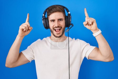 Foto de Hombre hispano con barba escuchando música con auriculares sonriendo asombrado y sorprendido y señalando con los dedos y los brazos levantados. - Imagen libre de derechos