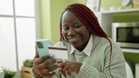 Téléchargez les photos : Femme africaine avec des cheveux tressés en utilisant smartphone et carte de crédit à la salle à manger - en image libre de droit