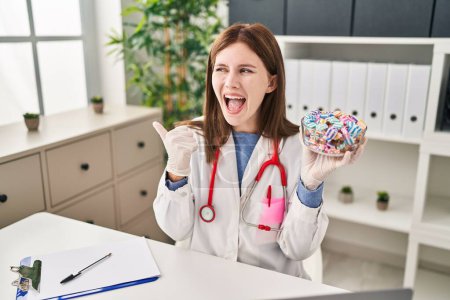 Photo for Young doctor woman holding sweets candy pointing thumb up to the side smiling happy with open mouth - Royalty Free Image