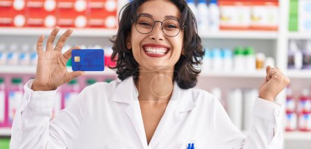 Foto de Joven mujer hispana trabajando en farmacia con tarjeta de crédito gritando orgullosa, celebrando la victoria y el éxito muy emocionada con el brazo levantado - Imagen libre de derechos