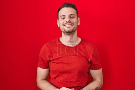 Foto de Young hispanic man standing over red background with hands together and crossed fingers smiling relaxed and cheerful. success and optimistic - Imagen libre de derechos