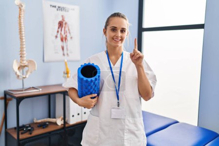 Photo for Young physiotherapist woman holding foam roll at the clinic surprised with an idea or question pointing finger with happy face, number one - Royalty Free Image