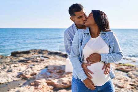 Téléchargez les photos : Jeune couple latin s'attendant bébé se câlinant et s'embrassant au bord de la mer - en image libre de droit