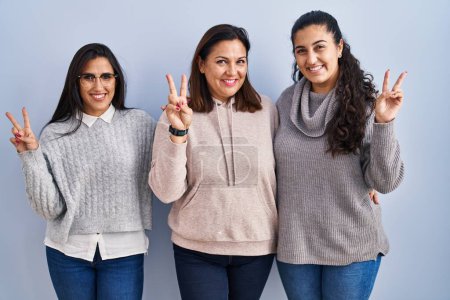 Photo for Mother and two daughters standing over blue background smiling with happy face winking at the camera doing victory sign with fingers. number two. - Royalty Free Image