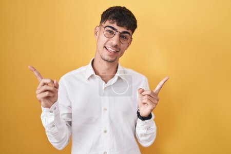Téléchargez les photos : Jeune homme hispanique debout sur fond jaune souriant confiant pointant avec les doigts vers différentes directions. espace de copie pour la publicité - en image libre de droit