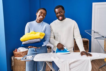 Téléchargez les photos : Jeune couple afro-américain repassage vêtements à la buanderie coller la langue heureux avec expression drôle. - en image libre de droit