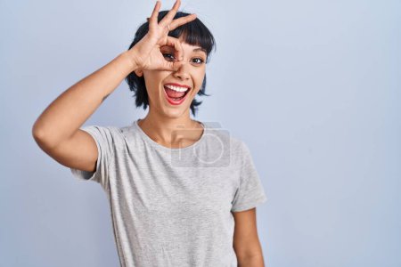 Foto de Mujer hispana joven vistiendo camiseta casual sobre fondo azul haciendo buen gesto con la mano sonriente, ojos mirando a través de los dedos con cara feliz. - Imagen libre de derechos