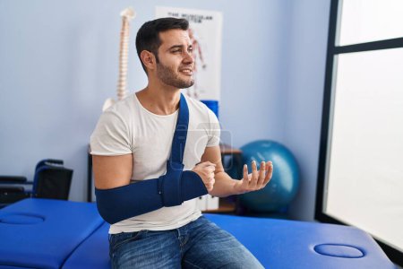 Foto de Hombre hispano joven lesionado paciente de fisioterapia que sufre de dolor en el brazo en la clínica de rehabilitación - Imagen libre de derechos