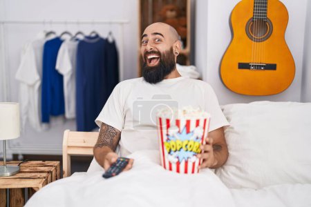 Téléchargez les photos : Jeune homme hispanique avec barbe et tatouages manger du pop-corn dans le lit célébrant fou et étonné pour le succès avec les yeux ouverts hurlant excité. - en image libre de droit
