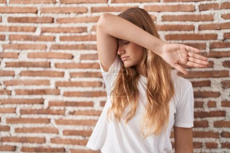 Photo for Young caucasian woman standing over bricks wall covering eyes with arm, looking serious and sad. sightless, hiding and rejection concept - Royalty Free Image