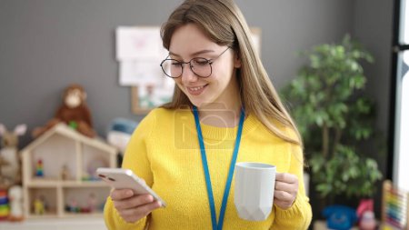 Téléchargez les photos : Young blonde woman preschool teacher using smartphone drinking coffee at kindergarten - en image libre de droit