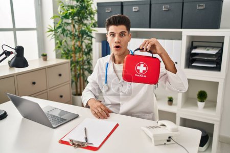 Photo for Young hispanic doctor man holding first aid kit scared and amazed with open mouth for surprise, disbelief face - Royalty Free Image