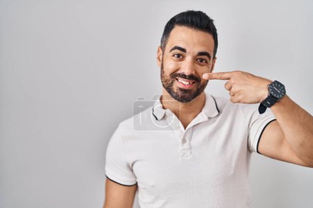 Téléchargez les photos : Jeune homme hispanique à la barbe portant des vêtements décontractés sur fond blanc pointant avec le doigt de la main sur le visage et le nez, souriant gai. concept de beauté - en image libre de droit