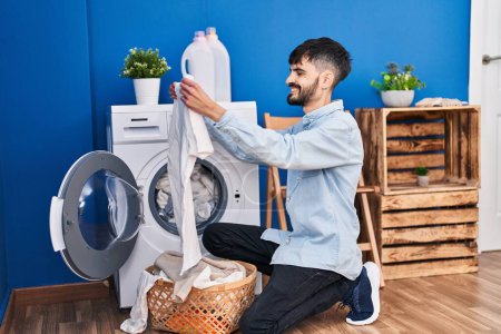 Foto de Young hispanic man holding white t shirt washing clothes at laundry room - Imagen libre de derechos