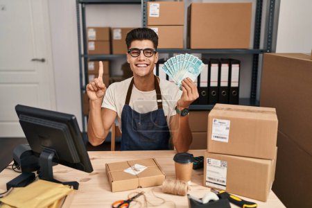 Photo for Young hispanic man working at small business ecommerce holding brazilian reals smiling with an idea or question pointing finger with happy face, number one - Royalty Free Image