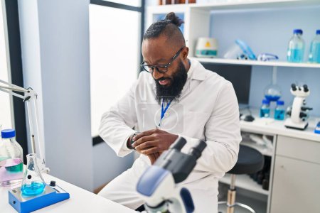 Foto de Hombre afroamericano joven vistiendo uniforme científico mirando reloj en el laboratorio - Imagen libre de derechos