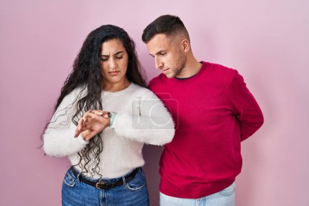Photo for Young hispanic couple standing over pink background checking the time on wrist watch, relaxed and confident - Royalty Free Image