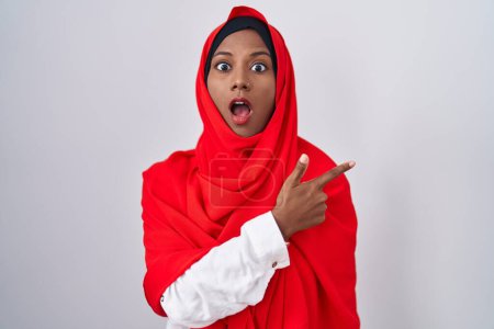 Foto de Mujer árabe joven con bufanda islámica tradicional hijab sorprendida señalando con el dedo hacia un lado, boca abierta expresión asombrada. - Imagen libre de derechos