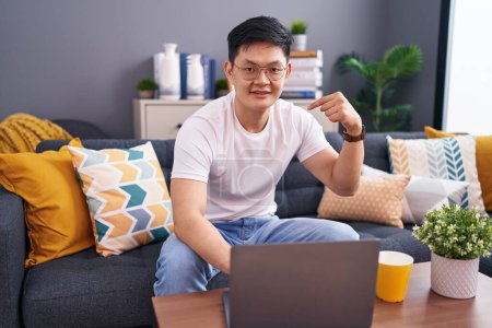 Foto de Joven asiático hombre usando laptop en casa sentado en el sofá buscando confidente con sonrisa en la cara, apuntando uno mismo con los dedos orgulloso y feliz. - Imagen libre de derechos