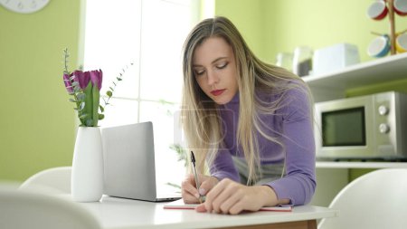 Photo for Young blonde woman writing on notebook using laptop at home - Royalty Free Image