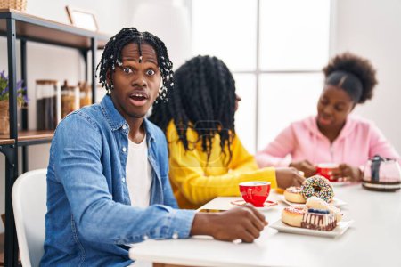 Photo for Group of three young black people sitting on a table having coffee scared and amazed with open mouth for surprise, disbelief face - Royalty Free Image