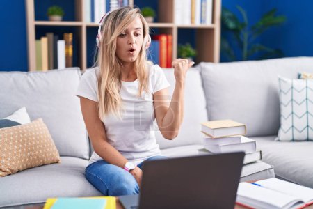 Photo for Young blonde woman studying using computer laptop at home surprised pointing with hand finger to the side, open mouth amazed expression. - Royalty Free Image