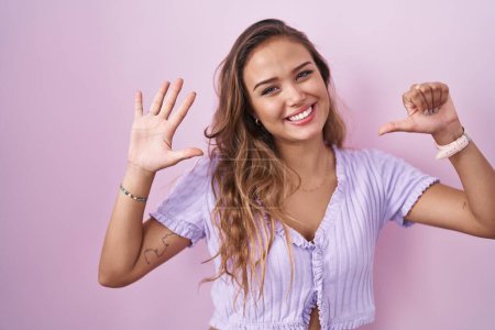 Foto de Mujer hispana joven de pie sobre fondo rosa mostrando y señalando hacia arriba con los dedos número seis mientras sonríe confiado y feliz. - Imagen libre de derechos