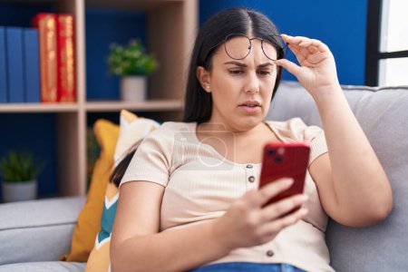 Téléchargez les photos : Jeune belle femme hispanique regardant smartphone écran avec problème de vision à la maison - en image libre de droit