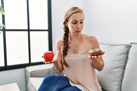 Foto de Mujer joven caucásica eligiendo comida saludable o poco saludable sentada en el sofá en casa - Imagen libre de derechos