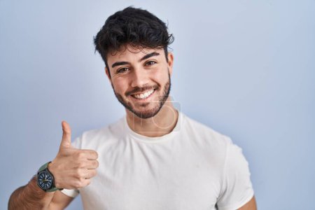 Foto de Hispanic man with beard standing over white background doing happy thumbs up gesture with hand. approving expression looking at the camera showing success. - Imagen libre de derechos