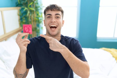 Téléchargez les photos : Jeune homme hispanique assis sur le lit tenant préservatif souriant heureux pointant avec la main et le doigt - en image libre de droit