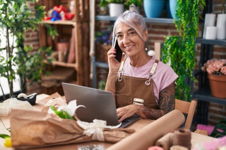 Photo for Middle age grey-haired woman florist talking on smartphone using laptop at florist - Royalty Free Image