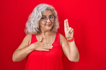 Foto de Mujer de mediana edad con el pelo gris de pie sobre el fondo rojo sonriendo jurando con la mano en el pecho y los dedos hacia arriba, haciendo una promesa de lealtad juramento - Imagen libre de derechos