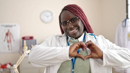 Téléchargez les photos : Femme africaine avec cheveux tressés médecin souriant faire geste de coeur avec les mains à la clinique - en image libre de droit