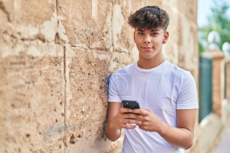 Téléchargez les photos : Jeune adolescent hispanique souriant confiant en utilisant un smartphone dans la rue - en image libre de droit