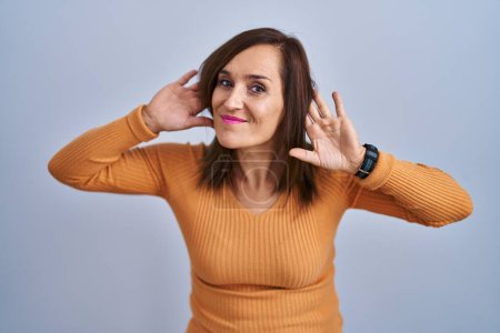 Téléchargez les photos : Femme brune du moyen âge portant un pull orange essayant d'entendre les deux mains sur le geste de l'oreille, curieuse de commérages. problème auditif, sourd - en image libre de droit