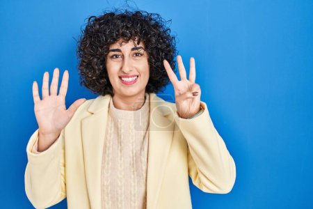 Téléchargez les photos : Jeune femme brune avec les cheveux bouclés debout sur fond bleu montrant et pointant vers le haut avec les doigts numéro huit tout en souriant confiant et heureux. - en image libre de droit