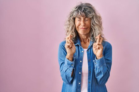 Téléchargez les photos : Femme d'âge moyen debout sur fond rose geste doigt croisé souriant avec espoir et les yeux fermés. chance et concept superstitieux. - en image libre de droit