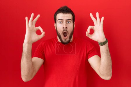 Photo for Young hispanic man wearing casual red t shirt looking surprised and shocked doing ok approval symbol with fingers. crazy expression - Royalty Free Image