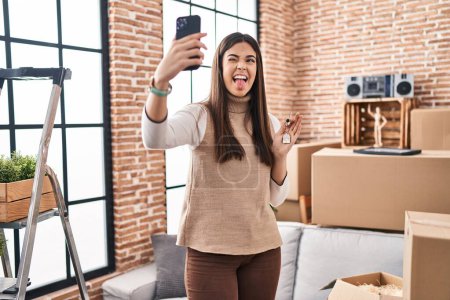 Photo for Young brunette woman moving to a new home doing video call sticking tongue out happy with funny expression. - Royalty Free Image