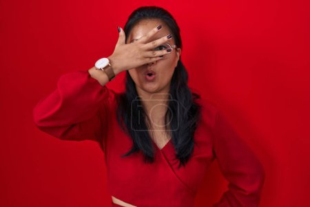 Téléchargez les photos : Asiatique jeune femme debout sur fond rouge regardant dans le choc couvrant le visage et les yeux avec la main, regardant à travers les doigts avec une expression embarrassée. - en image libre de droit