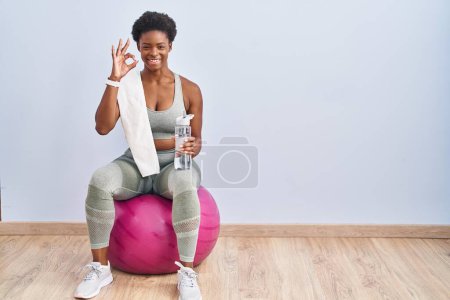 Photo for African american woman wearing sportswear sitting on pilates ball smiling positive doing ok sign with hand and fingers. successful expression. - Royalty Free Image