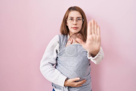 Foto de Mujer caucásica joven sosteniendo y llevando al bebé en un cabestrillo con la mano abierta haciendo señal de stop con expresión seria y segura, gesto de defensa - Imagen libre de derechos