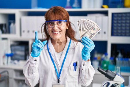 Téléchargez les photos : Femme hispanique du Moyen Âge travaillant au laboratoire scientifique tenant des dollars surpris par une idée ou une question pointant du doigt avec un visage heureux, numéro un - en image libre de droit