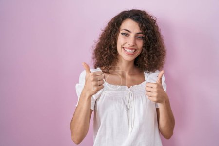Foto de Mujer hispana con el pelo rizado de pie sobre el signo de éxito de fondo rosa haciendo gesto positivo con la mano, pulgares hacia arriba sonriendo y feliz. expresión alegre y gesto ganador. - Imagen libre de derechos