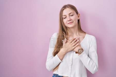 Foto de Young caucasian woman standing over pink background smiling with hands on chest with closed eyes and grateful gesture on face. health concept. - Imagen libre de derechos